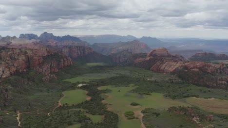 High-and-wide-slow-panoramic-drone-shot-of-Zion-Nation-Park-valley,-green-grass-and-prominent-red-rocks