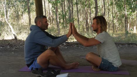 Two-Men-Sitting-On-The-Yoga-Mat-Facing-Each-Other-Doing-Eye-To-Eye---Palm-To-Palm-As-Connection---Queensland,-Australia