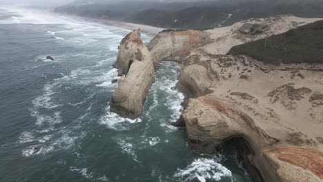 Two-paragliders-flying-over-Cape-Kiwanda-rugged-rock-formation-heavily-eroded-by-the-ocean,-aerial