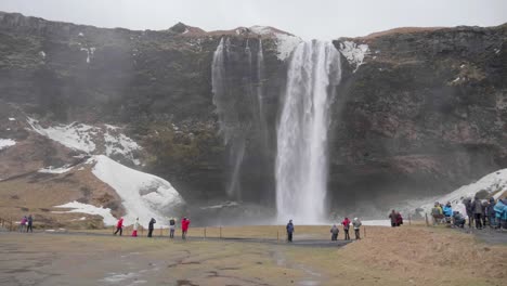 Static,-slow-motion-shot,-of-lots-of-people-standing-in-front-and-enjoying-the-view-of-seljalandfoss-waterfall,-on-a-cloudy,-autumn-day,-in-Iceland