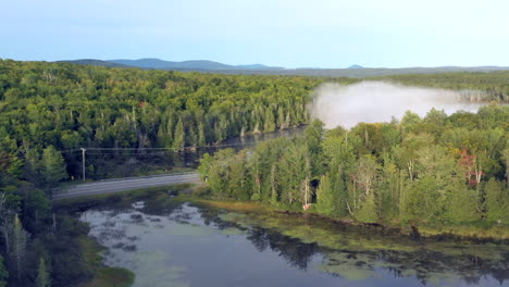 Aerial-drone-shot-emerging-from-thick-fog-and-mist-to-reveal-the-blue-waters-of-Spectacle-Pond-and-a-road-running-through-the-green-forest-of-the-Maine-wilderness