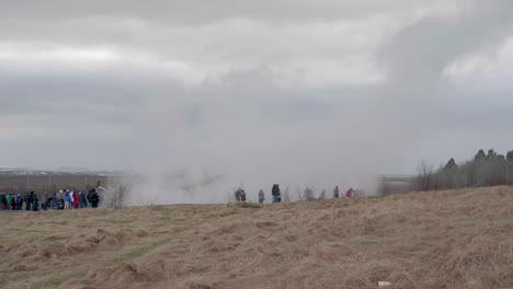 Static,-wide-shot,-of-lots-of-people-standing-in-front-of-the-erupting-geisir,-on-a-cloudy,-autumn-day,-in-Geysir,-Iceland