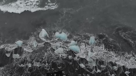 Slow-motion-panning-drone-shot-of-waves-crashing-into-black-sand-beach-icebergs