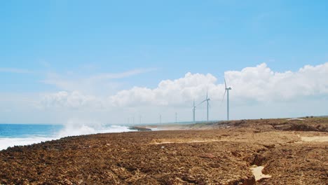 Wind-turbines-spinning-along-rocky-beach-with-ocean-waves-in-Curacao,-Caribbean