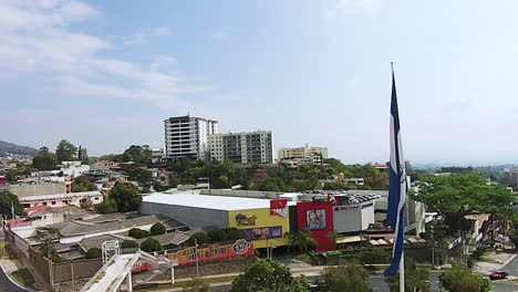 Drone-shot-of-the-Salvadoran-flag-blowing-in-the-wind-while-overlooking-the-city-skyline
