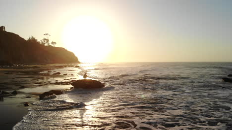 Ein-Mann-In-Silhouette-Bei-Sonnenaufgang-Beobachtet-Mit-Einer-Luftdrohne,-Wie-Die-Meereswellen-Am-Strand-Aufschlagen,-Und-Zieht-Sich-Zurück,-Um-Die-Atemberaubende-Küste-Von-Santa-Barbara,-Kalifornien,-Zu-Betrachten