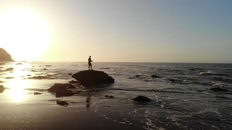 Ein-Mann-In-Silhouette-Bei-Sonnenaufgang,-Der-Beobachtet,-Wie-Die-Meereswellen-Am-Strand-An-Der-Küste-Von-Santa-Barbara,-Kalifornien,-Aus-Der-Luft-Krachen