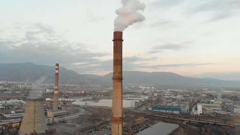 Industrial-chimney-smoking,-buildings-and-mountains-as-background---aerial-shot