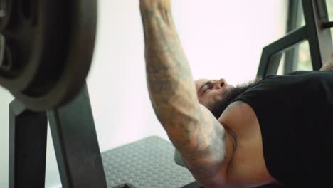 Attractive-sporty-man-with-tattoos-and-beard-at-training-bench-press-in-gym-close-up