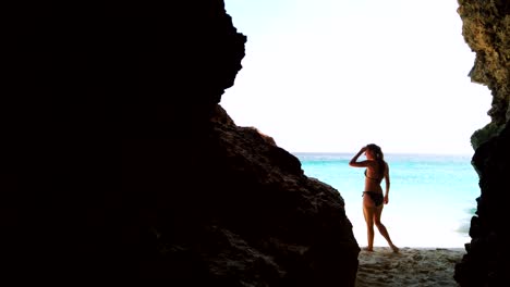 Eine-Halbsilhouette-Aufnahme-Einer-Jungen-Frau,-Die-Eine-Dunkle-Höhle-Verlässt-Und-Zu-Einem-Sonnigen-Strand-Mit-Weißem-Sand-Und-Kristallklarem-Wasser-Geht