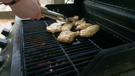 Hand-with-tongs-reaches-in-to-flip-half-cooked-chicken-over-on-the-grill
