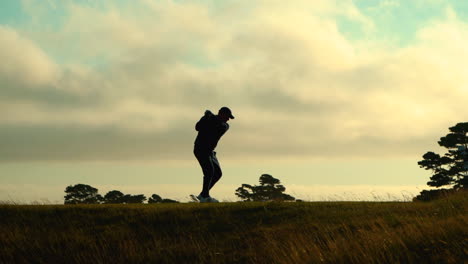 Silhouetted-golfer-taking-a-swing-and-a-second-player-walks-up,-beautiful-clouds-and-sky,-Bandon-Dunes-Golf-Resort