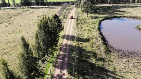 Aerial-video-of-a-car-leaving-a-house-in-the-countryside-while-a-dog-runs-it,-with-a-lake-in-the-background-and-many-trees
