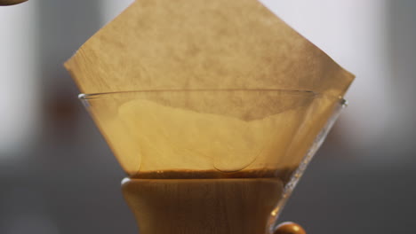 Close-up-view-of-kettle-pouring-hot-water-into-large-glass-Chemex-coffee-maker-over-fresh,-ground-coffee-beans-in-the-morning
