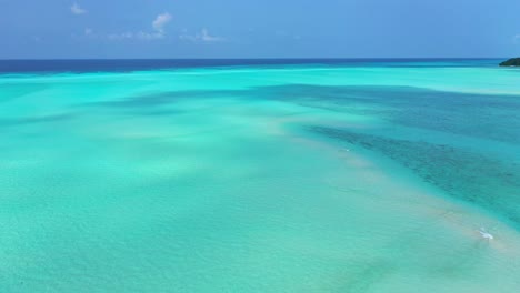 Turquoise-sea-texture-with-calm-crystal-water-over-white-sandy-bottom-bordered-by-deep-blue-ocean-and-bright-sky,-Maldives