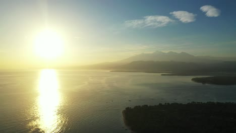 Sunset-over-ocean-horizon,-glowing-yellow-sky-reflecting-on-calm-sea-surface-washing-shore-of-tropical-islands,-Bali