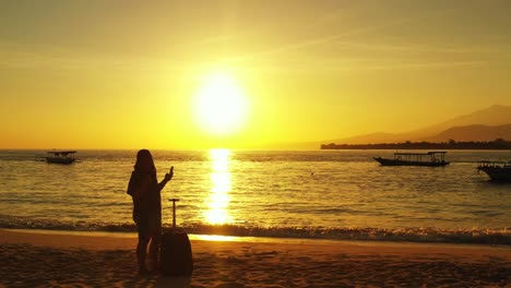 Silhouette-of-a-young-woman-with-a-piece-of-luggage-taking-selfies-on-the-paradise-sandy-beach-during-the-golden-sunset
