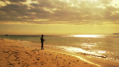 Young-man-fishing-during-a-beautiful-orange-and-yellow-sunset-on-a-beach-paradise