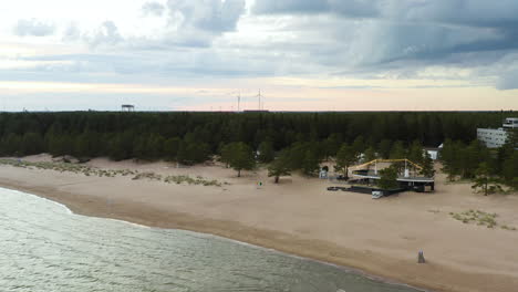 Aerial-view-of-a-restaurant-on-the-Yyteri-beach,-rising-and-revealing-windmills-and-and-colorful,-cloudy-dusk-sky,-in-Pori,-Finland---Crane,-drone-shot