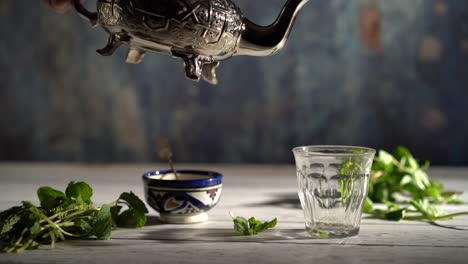Slow-motion-wide-shot-of-hand-picking-up-an-ornate-teapot-and-pours-tea-into-a-glass-with-Moroccan-bowl-and-mint-around