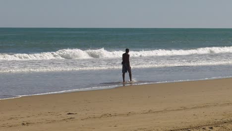 Tall-man-walks-in-shallow-waves-on-sandy-shore-in-summertime,-slow-motion