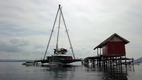 Trimaran-sailboat-Bigkanu-docked-on-an-ocean-reef-dive-cottage-painted-red,-Aerial-circle-around-shot