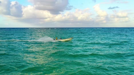Small-narrow-motor-boat-carrying-three-people-travels-on-green-ocean-sea-waters,-Punta-Cana,-Dominican-Republic,-circle-aerial-approach