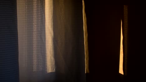 A-curtain-is-moved-slowly-by-the-breeze,-with-warm-dusk-light-falling-on-a-nearby-wall-fading-slowly-to-black