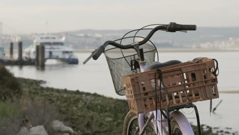 Vieja-Bicicleta-Con-Canasta-Estacionada-En-La-Playa-Con-Pasajeros-Entrando-En-Un-Ferry-En-Segundo-Plano