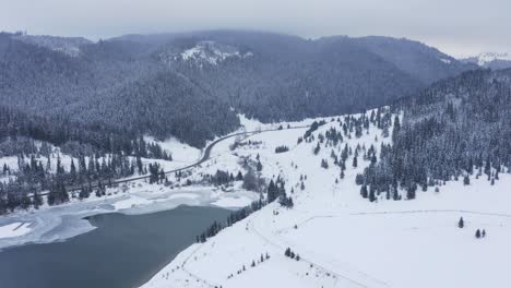 Cars-drive-on-windy-mountain-valley-road-in-frozen-winter-snow-landscape,-drone-crane-shot