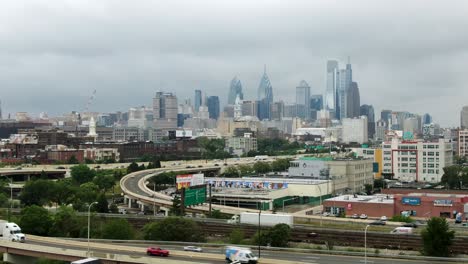 Aerial-establishing-shot-of-Philadelphia-skyline,-traffic-flowing-with-skyscrapers-in-distance-on-cloudy-summer-day