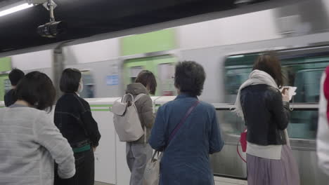 Viajeros-De-Pie-En-La-Plataforma-De-La-Estación-De-Metro-Con-Un-Tren-Que-Pasa-Por-Tokio,-Japón