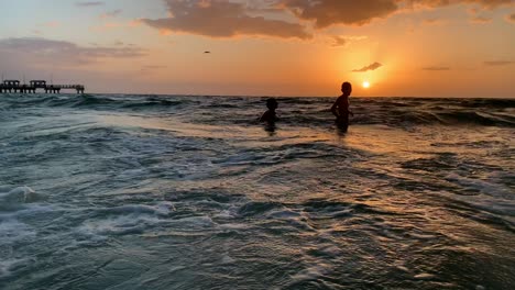 Dos-Niños-Nadando---Saltando-Olas-En-El-Océano-En-La-Playa-Durante-La-Puesta-De-Sol-En-Florida