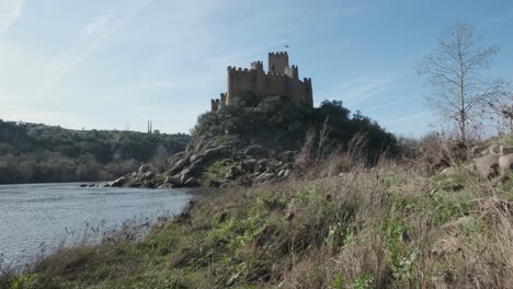 Spectacular-scenic-view-of-medieval-Castle-of-Almourol-atop-islet-in-middle-of-Tagus-River-water,-Praia-do-Ribatejo,-on-sunny-blue-sky-day,-Portugal,-handheld-pan-up