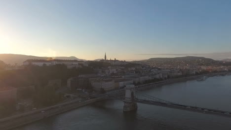 Aerial-shot-of-the-Chain-bridge,-Panning-left-from-Pest,-to-Buda-side-showing-the-sunset-over-the-castle