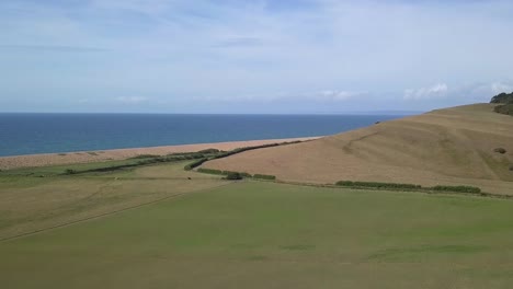 Low-forward-tracking-aerial-moving-upwards-over-a-field-revealing-the-vast-expanse-of-the-dorset-coastline-at-the-west-end-of-Chesil-Beach
