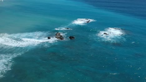 Wunderschöne-Luftaufnahme-Der-Vielen-Schattierungen-Des-Klaren-Blauen-Wassers-Der-Karibik,-Während-Sich-Die-Wellen-über-Große-Felsen-Brechen