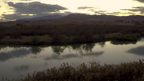 Zeitrafferschwenk-Eines-Sonnenuntergangs,-Der-Von-Der-Glasigen-Oberfläche-Eines-Flusses-Mit-Bergen-Im-Hintergrund-Reflektiert-Wird