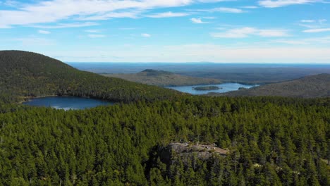 Aerial-drone-establishing-shot-over-the-vast-green-and-autumn-colored-forest-with-blue-lakes-dotting-the-wilderness-and-a-large-group-of-hikers-on-a-rocky-outcrop-in-Maine