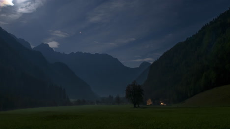 Timelapse-of-Alpine-valley-by-night-under-full-moon,-illuminated-farmhouse,-cottage,-full-moon,-clouds-and-stars-in-sky,-zoom-out-pull,-revealing-shot