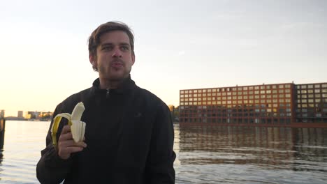 Handsome-man-eating-a-banana-and-smiling-on-a-pier-during-sunset