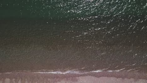 Aerial-top-shot-of-shimmering-waves-washing-onto-a-pebble-beach,-STATIC-CROP