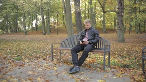 Ein-Junger-Attraktiver-Mann-Sitzt-An-Einem-Herbsttag-Auf-Einer-Parkbank-Und-Benutzt-Sein-Smartphone