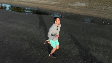 Aerial-tracking-shot-of-fit-young-man-running-on-a-beach-in-Auckland,-New-Zealand