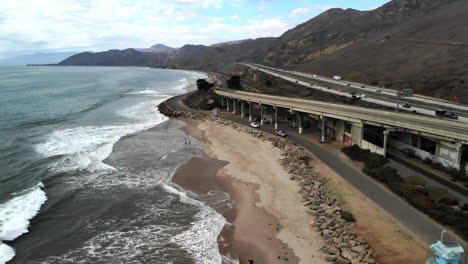 Aerial-drone-shot-orbiting-around-a-concrete-bridge-on-the-beach-in-Ventura-with-surfers-in-the-ocean-and-RV-campers-next-to-the-California-101-freeway-along-the-coast