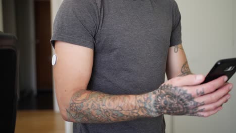 A-tattooed-young-man-with-diabetes-uses-his-phone-to-check-his-blood-glucose-levels-with-glucose-reading-sensor-on-his-arm-and-then-watches-the-info-on-the-phone