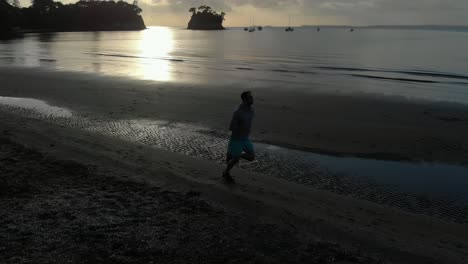 Aerial-shot-tracking-a-silhouetted-young-man-running-on-a-beach-in-Auckland,-New-Zealand