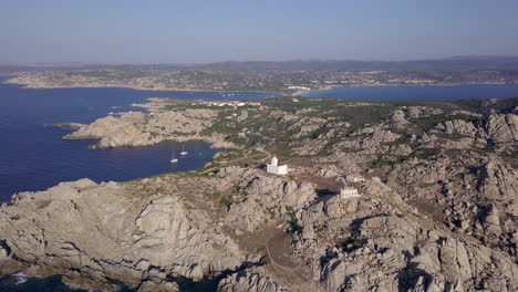 Drone-shot-orbiting-a-small-white-light-house-to-reveal-a-rocky-cliff-and-wild-coastline-in-north-Sardinia