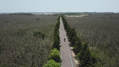 drone-shot-of-scooter-riding-through-a-single-road-pass-by-forest-trees