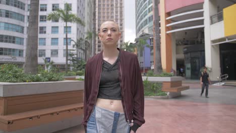A-woman-with-a-shaved-head-wearing-a-jacket-walks-past-stores-in-an-outdoor-mall-as-others-shop